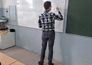 Prowadzący zajęcia stoi tyłem do uczniów i pisze na tablicy suchościeralnej.